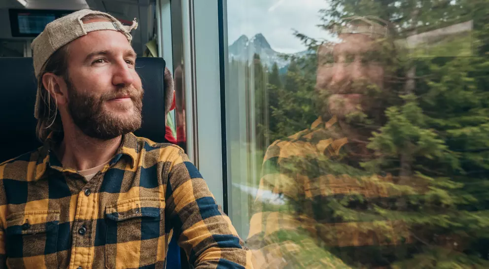 A man in a plaid shirt and cap gazes out the train window, with mountains and trees reflected in the glass.