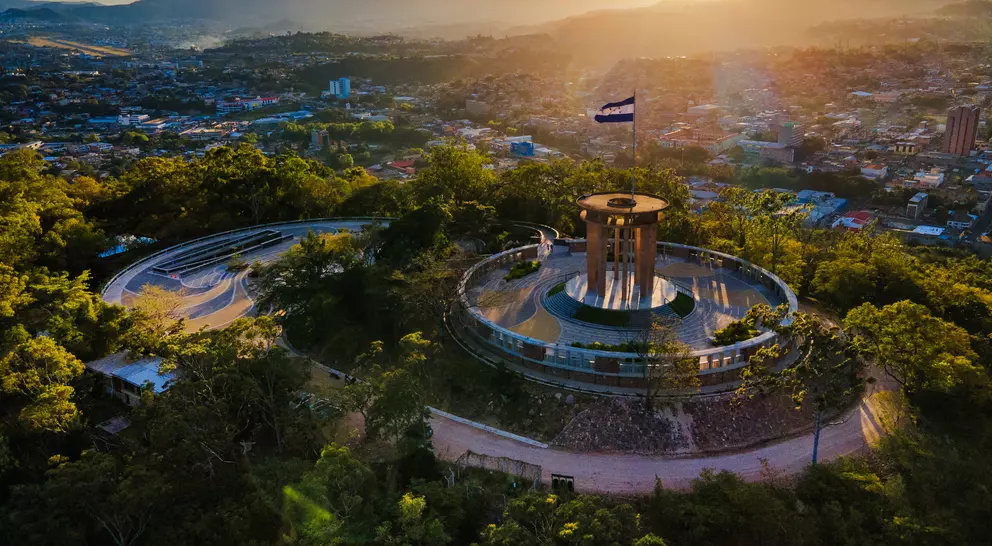 Aerial view of a scenic lookout with circular paths, a monument, and a flag, surrounded by trees and a city backdrop at sunset.