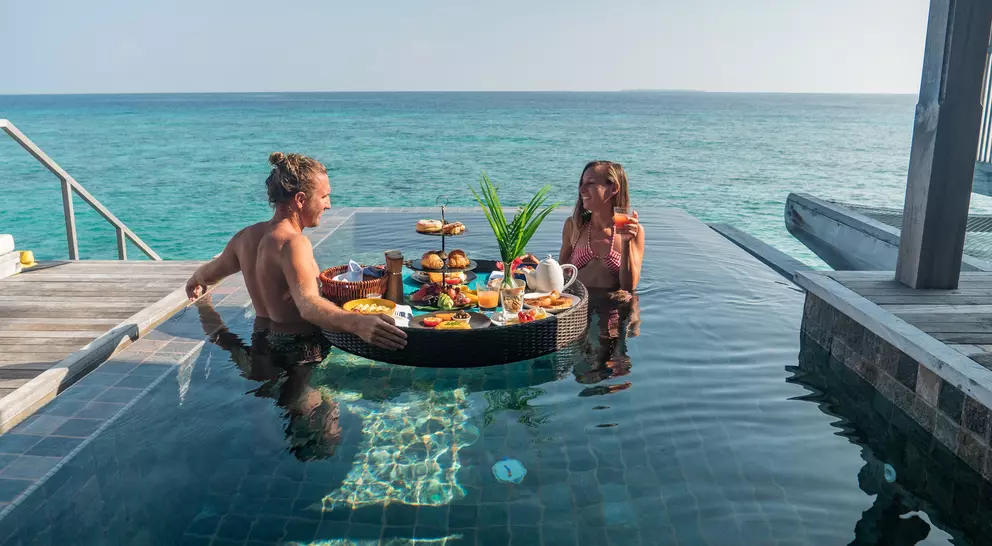 A couple relaxes in a pool, enjoying a floating breakfast with scenic ocean views.