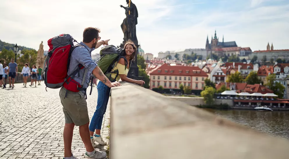 Two travelers with backpacks enjoy the view from a bridge, with historic buildings and a statue in the background.