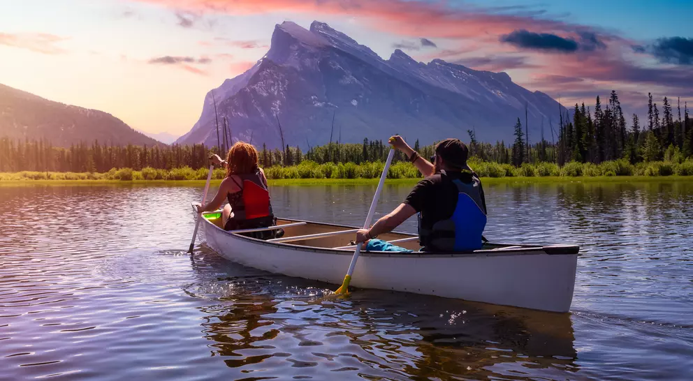 Two people paddle a canoe on a calm lake, with mountains and colorful sunset in the background.