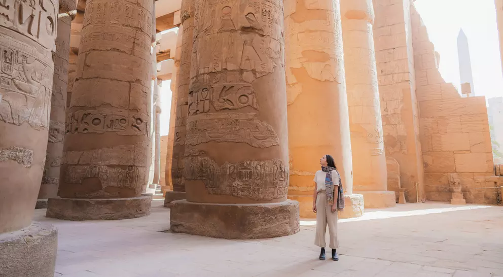 Person standing among tall, ancient, carved stone columns inside a historic site, with sunlight streaming in.