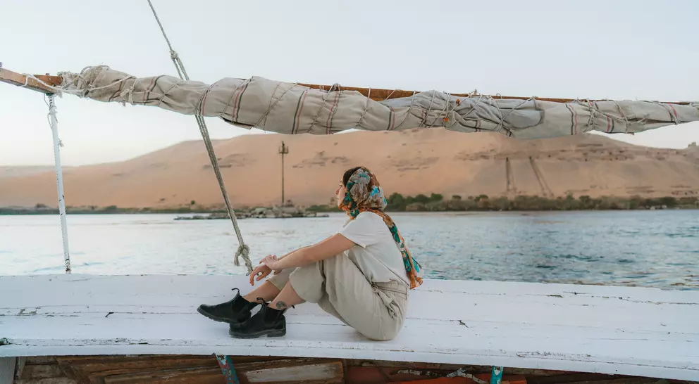 Person sitting on a boat, wearing a scarf, with desert hills and water in the background during sunset.