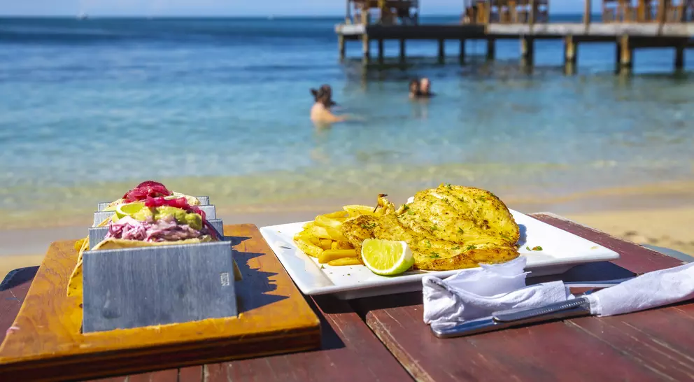 A beachside table displays two dishes: a colorful platter and a grilled fish dish, with people swimming in the background.