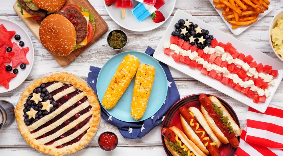A variety of festive foods including burgers, hot dogs, watermelon, corn, and desserts arranged in a patriotic theme.