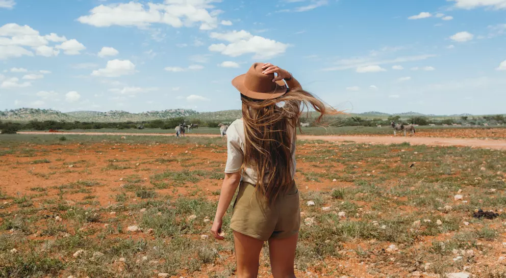 A person with long hair in a hat stands on a grassy plain, looking at a wide sky with scattered clouds.