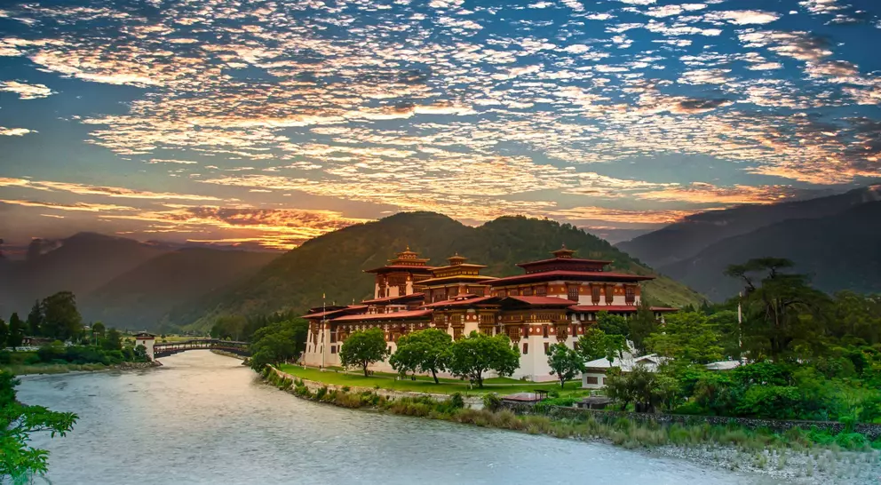 A scenic view of a traditional Bhutanese building by a river, surrounded by mountains and a vibrant sky at sunset.