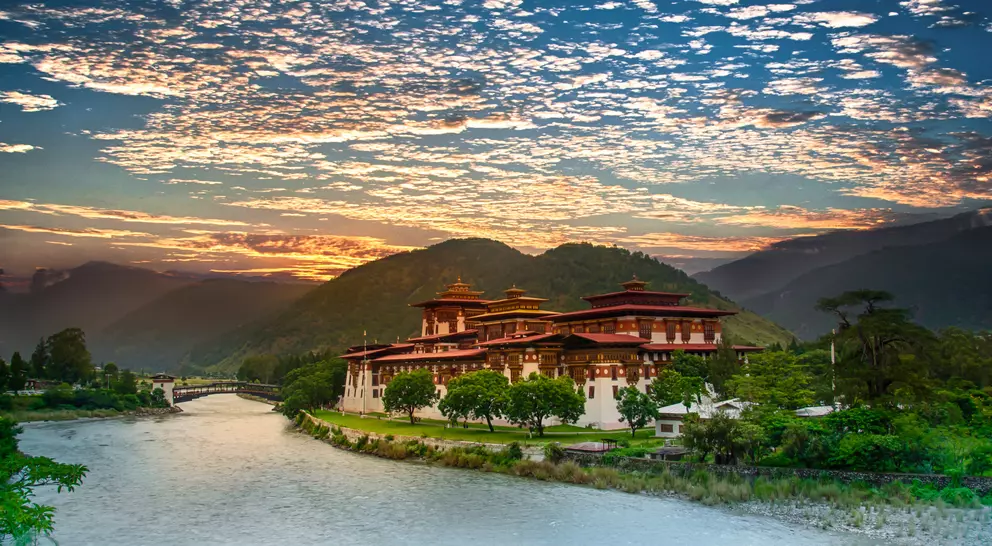 A scenic view of a traditional Bhutanese building by a river, surrounded by mountains and a vibrant sky at sunset.