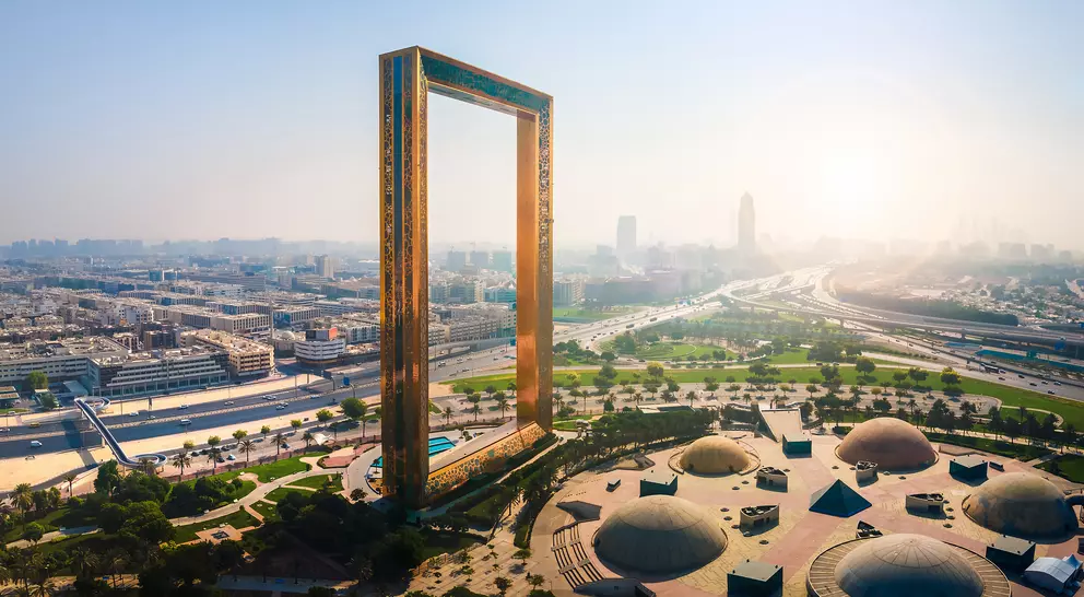 Aerial view of Dubai Frame, a large rectangular structure, with city skyline and green parks in the background.