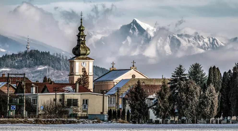 A snowy landscape with a church tower and mountains in the background, under a cloudy sky.