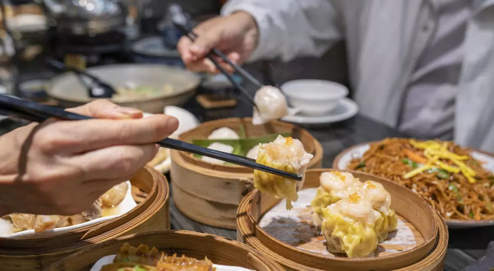 Hands using chopsticks to pick up dim sum from bamboo steamers on a table with noodles and other dishes in the background.