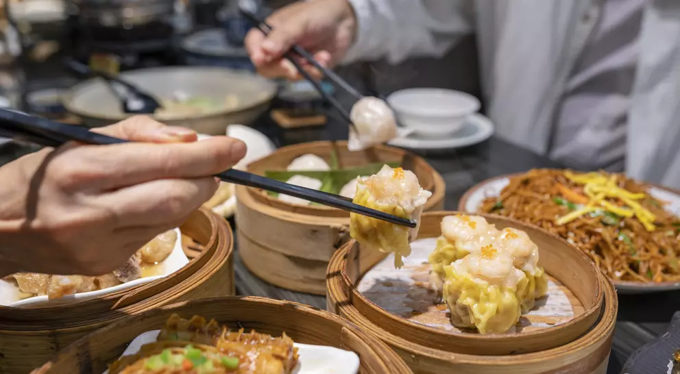 Hands using chopsticks to pick up dim sum from bamboo steamers on a table with noodles and other dishes in the background.