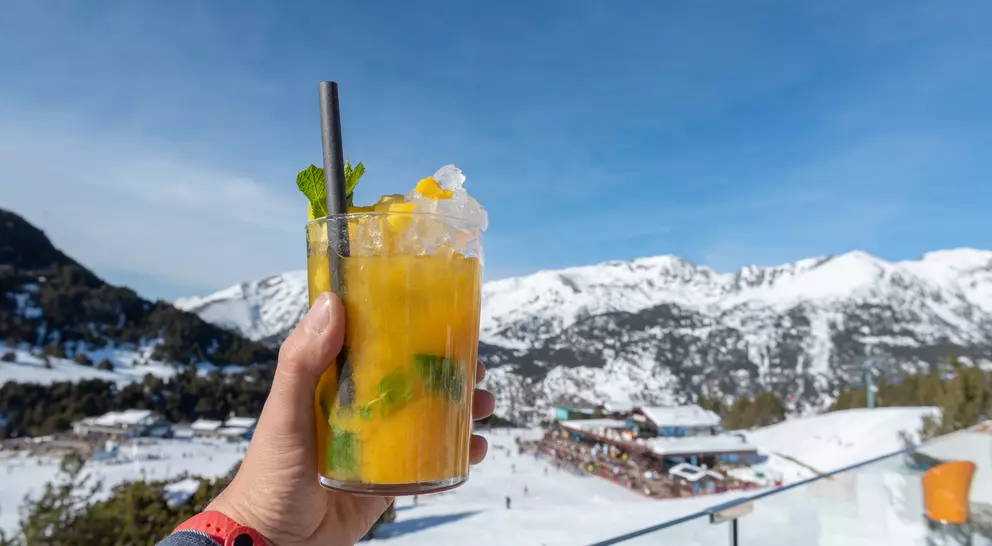 A hand holds a glass of orange cocktail with mint, set against a snowy mountain backdrop under a clear blue sky.