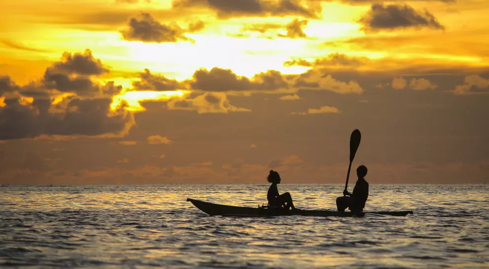 Two people sit in a kayak at sunset, silhouetted against a colorful sky with clouds reflecting in the water.
