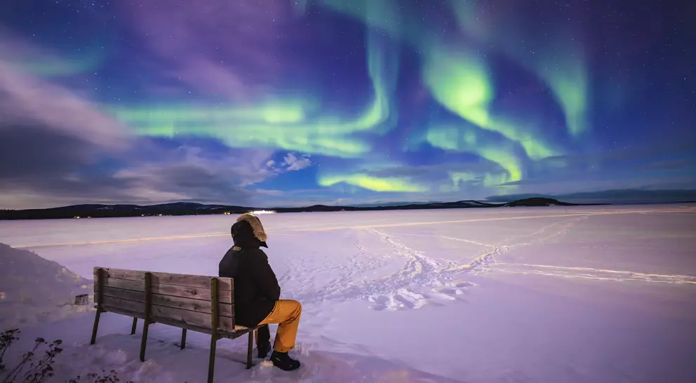 Person sitting on a bench, gazing at vibrant northern lights over a snowy landscape.