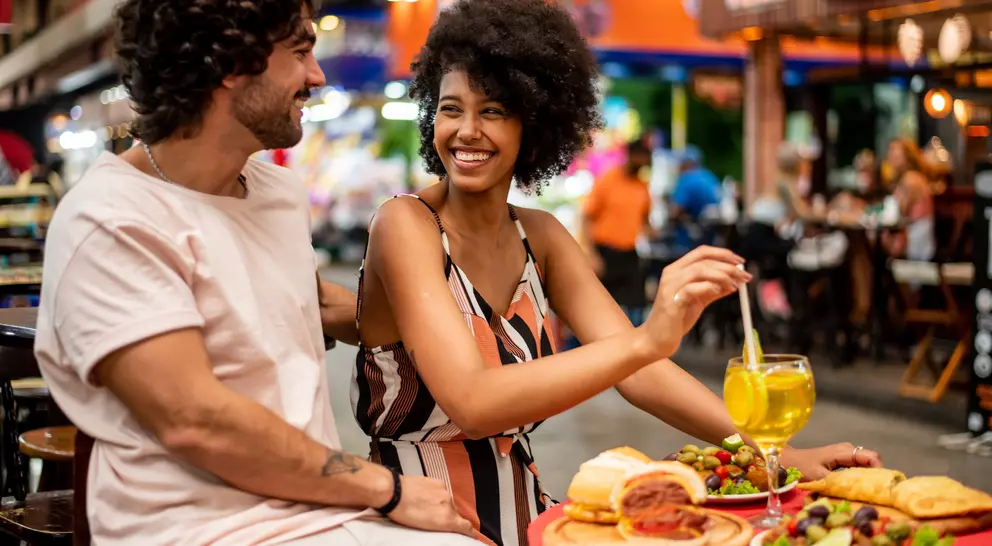 A couple enjoys a meal at a lively outdoor restaurant, smiling and sharing food and drinks in a vibrant atmosphere.