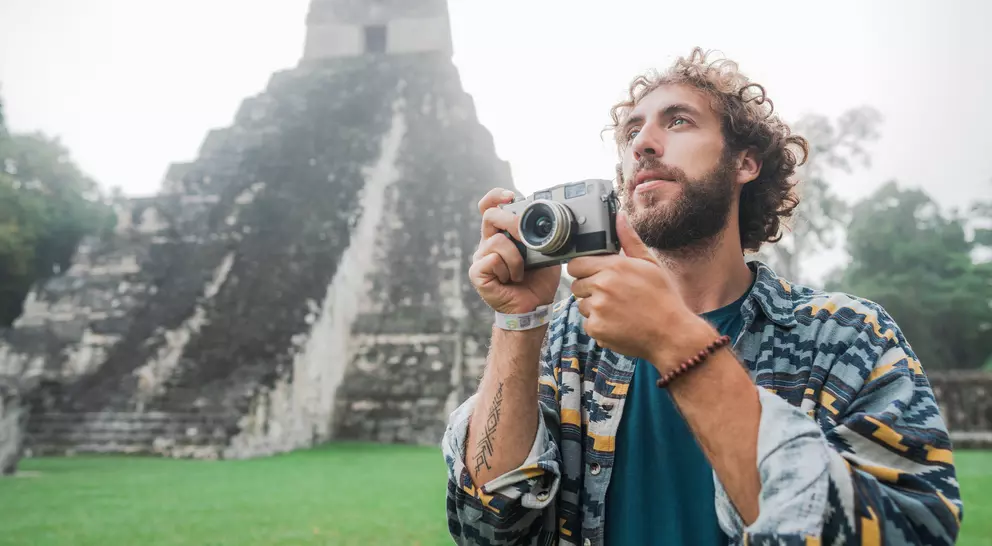 A man with curly hair holds a camera, standing in front of a Mayan pyramid in a misty landscape.