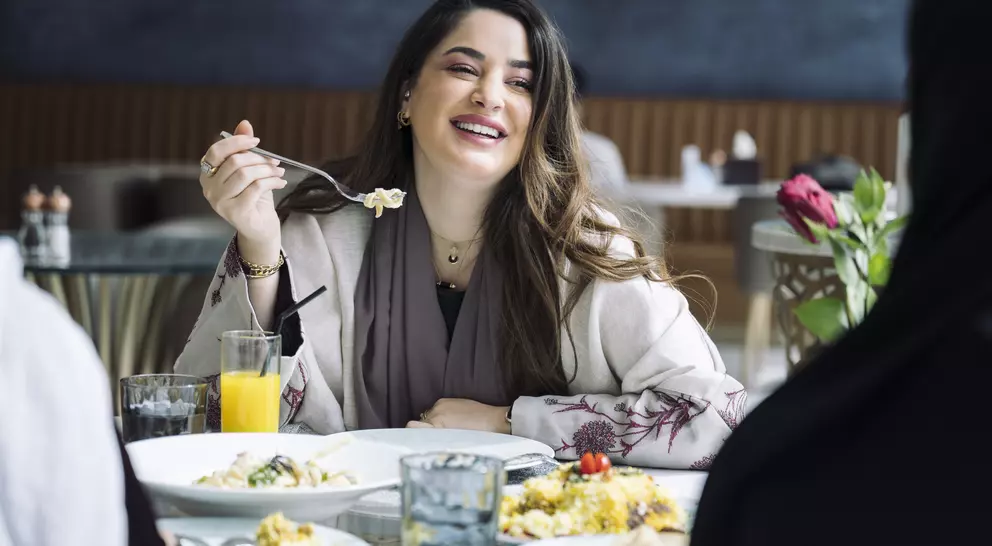 A woman smiles and enjoys a meal at a restaurant, surrounded by plates of food and drinks on the table.