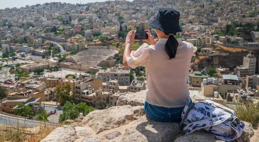 A person in a hat takes a photo of a city view, showcasing hills and buildings in the background.