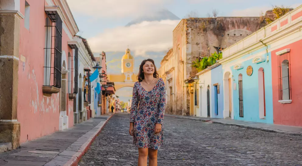 A woman stands on a cobblestone street lined with colorful buildings, with a mountain in the background under a clear sky.