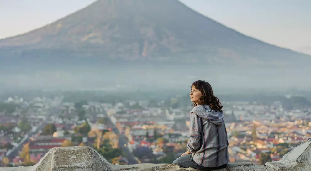 A person sits on a stone wall, overlooking a valley with a volcano in the background, surrounded by misty mountains.