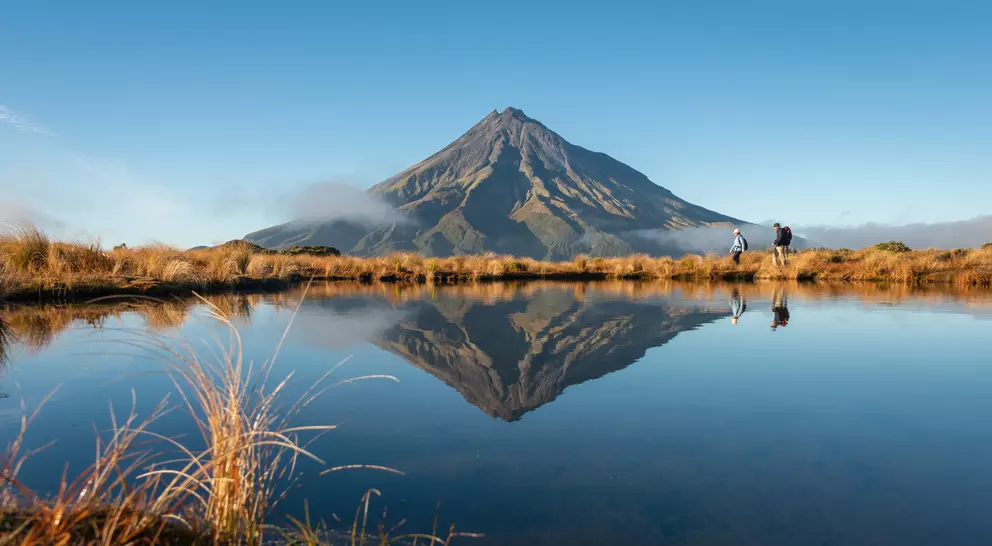A scenic view of a mountain reflected in a calm pond, with two hikers walking along the shore.