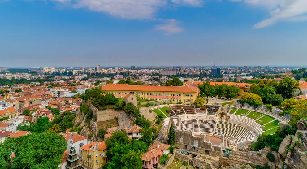 Aerial view of an ancient amphitheater surrounded by cityscape and greenery under a partly cloudy sky.