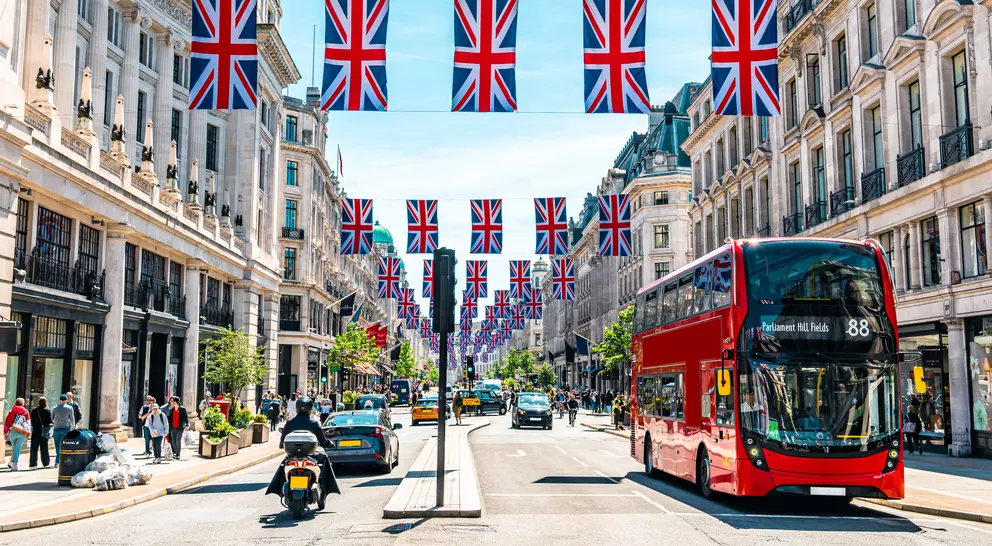 A busy street lined with shops, featuring red double-decker buses and decorative Union Jack flags overhead.