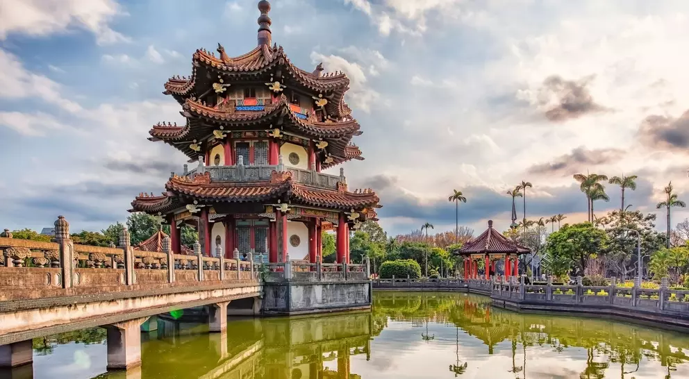 A traditional pagoda beside a tranquil pond, surrounded by lush greenery and a cloudy sky.