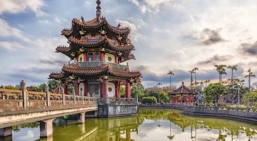 A traditional pagoda beside a tranquil pond, surrounded by lush greenery and a cloudy sky.
