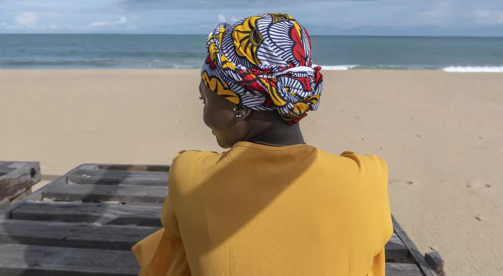 A woman wearing a colorful headwrap sits by the beach, facing the ocean, with a sandy shore in the background.