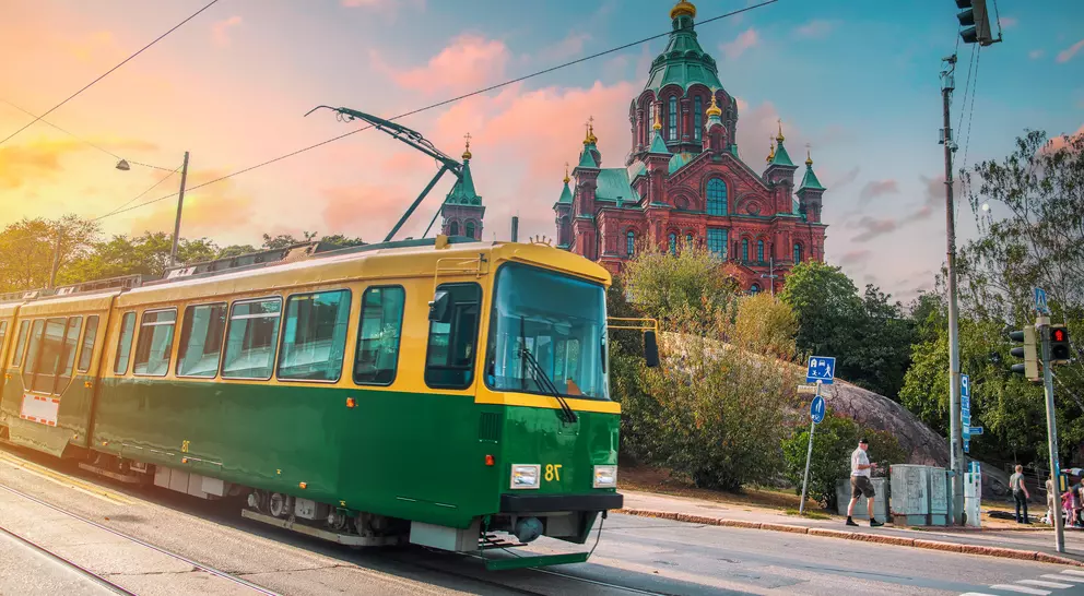 A green and yellow tram travels on a street with a historic building in the background at sunset.