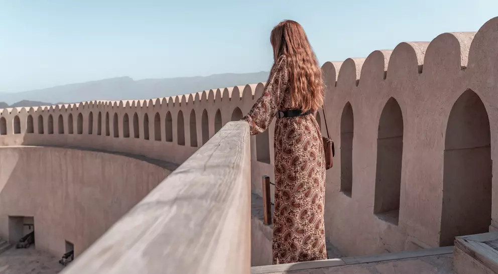 A woman in a patterned dress stands on a balcony, gazing at distant mountains under a clear blue sky.
