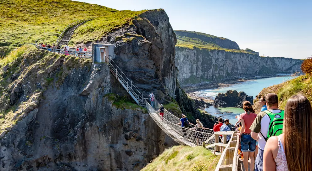 A sunny coastal scene showing a suspended bridge with people walking towards scenic cliffs and the ocean in the background.