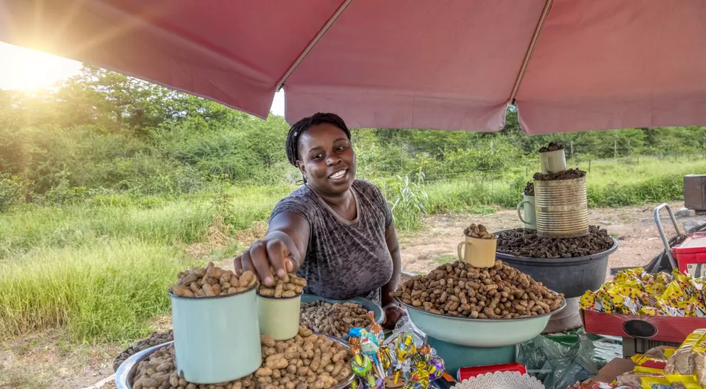 A smiling vendor at a market offers a cup of peanuts, surrounded by various snacks under a shaded tent.