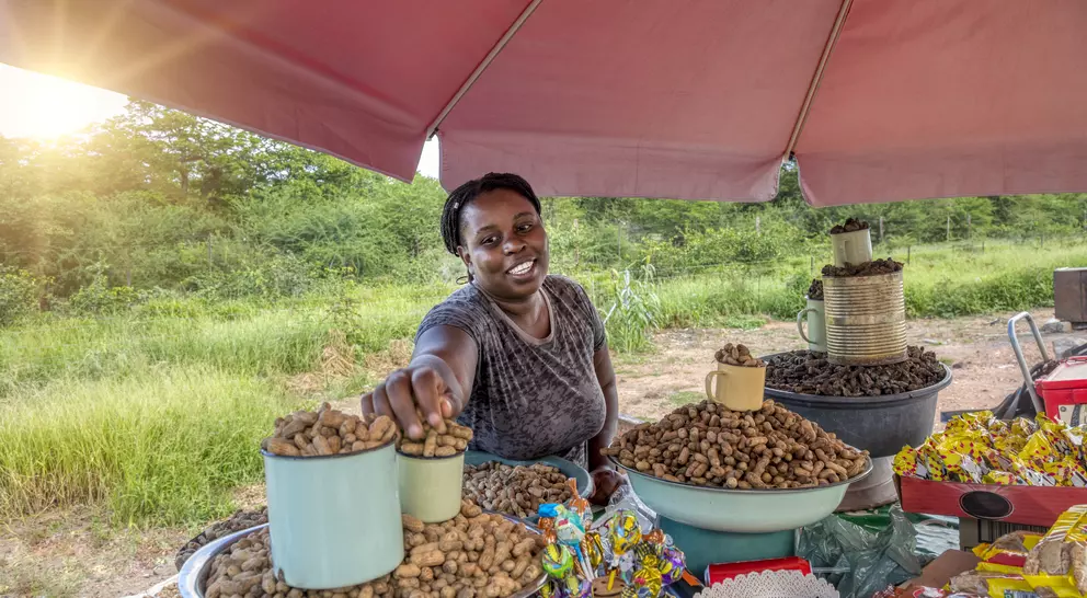 A smiling vendor at a market offers a cup of peanuts, surrounded by various snacks under a shaded tent.