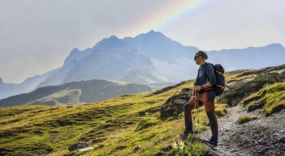 Hiker standing on a rocky trail, gazing at mountains and a rainbow in the sky.