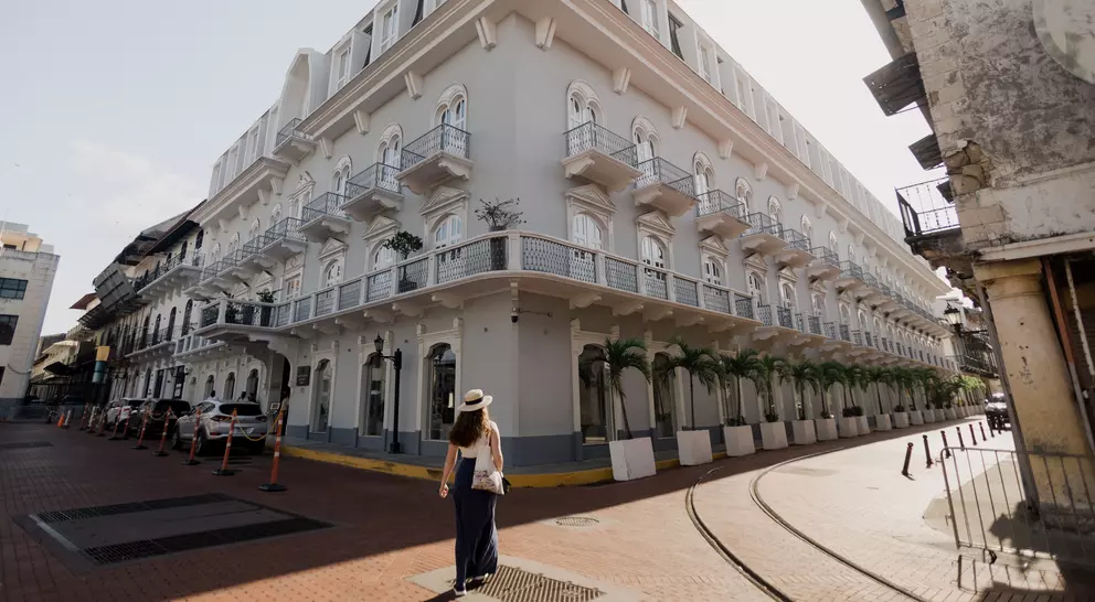 A woman walks along a cobblestone street beside a large, elegant white building with balconies and palm trees.