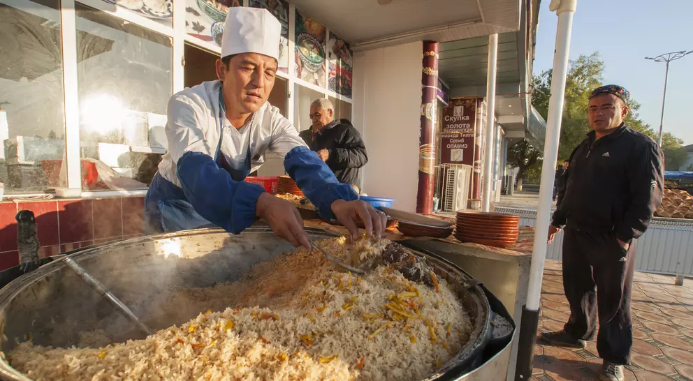 A chef in a white hat prepares a large pot of rice dish outdoors, while a man waits nearby.
