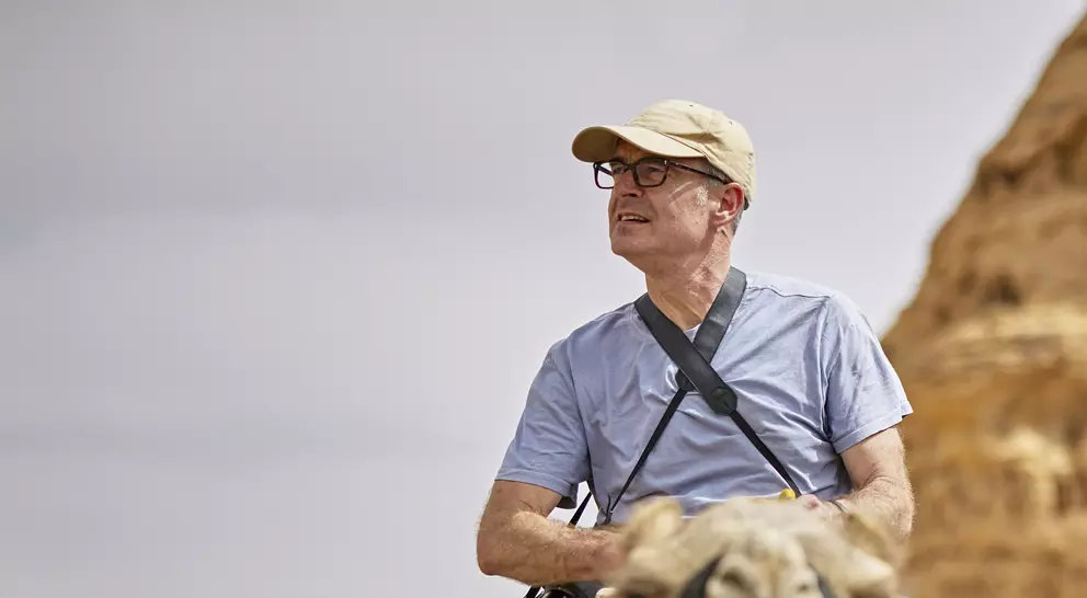 Two men ride camels in a desert landscape, with a large rock formation in the background. One man wears glasses and a cap.