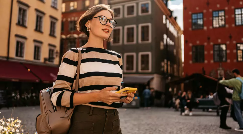 Young woman in a striped sweater stands in a plaza, holding a phone, with colorful buildings and people around her.