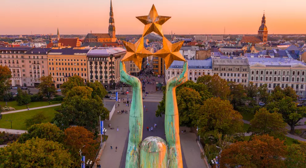 View of a city at sunset, featuring a monumental statue with raised hands holding three stars, surrounded by trees and buildings.