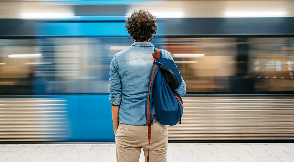 A person stands at a subway platform, facing a train speeding by, with a blue bag slung over their shoulder.