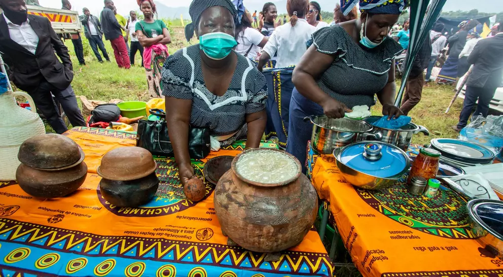 Two women prepare food outdoors at a vibrant market, with traditional dishes displayed on colorful patterned tablecloths.