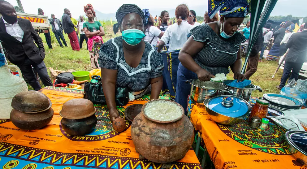 Two women prepare food outdoors at a vibrant market, with traditional dishes displayed on colorful patterned tablecloths.