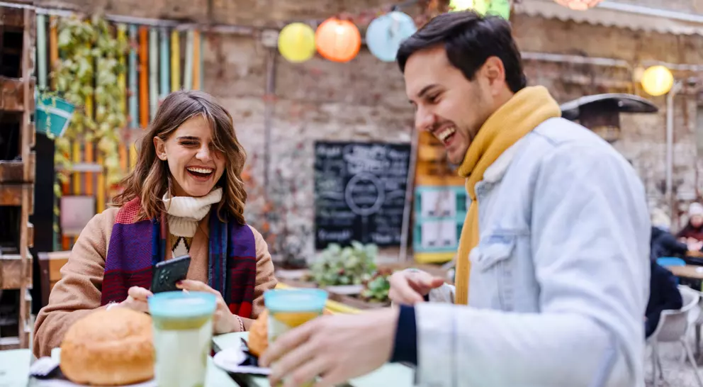 A man and woman sit at a colorful outdoor cafe, laughing and enjoying drinks and pastries.