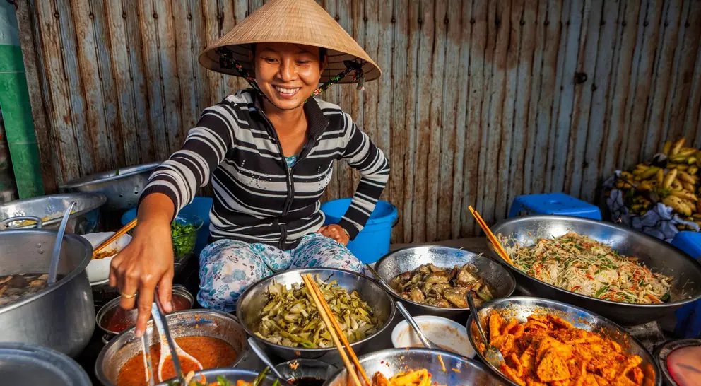 A smiling woman in a conical hat serves dishes from a variety of bowls at a vibrant market stall.