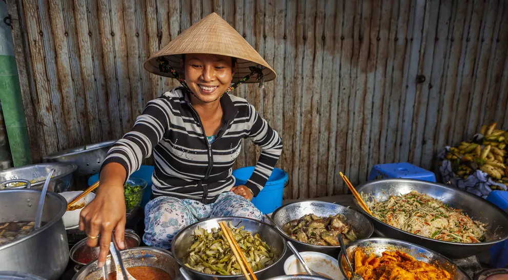 A smiling woman in a conical hat serves dishes from a variety of bowls at a vibrant market stall.