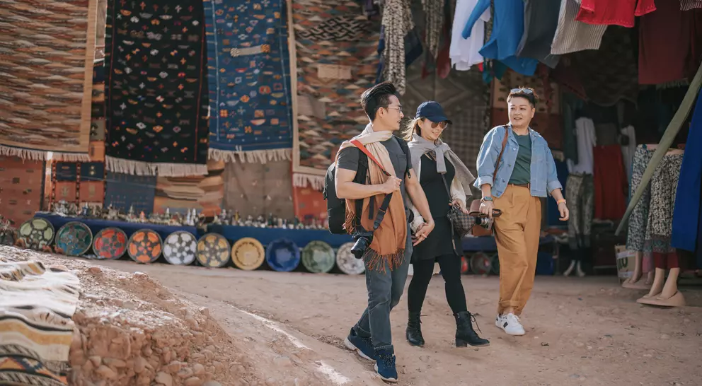 Three people walk through a vibrant market, surrounded by colorful textiles and handmade pottery.