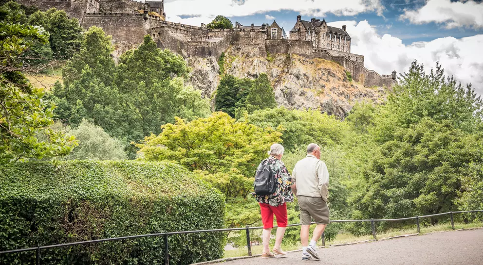 Two people stroll along a path, with Edinburgh Castle perched on a rocky hill in the background, surrounded by greenery.